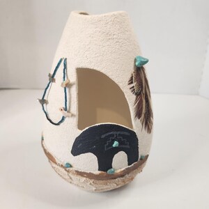May include: A cream-colored textured ceramic vase with a large side cutout. The vase features a painted black bear silhouette, a blue beaded strand, brown feathers, and small turquoise-colored stone accents near the base and top rim.