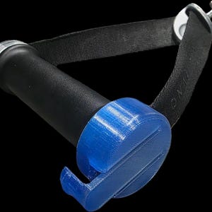 Può includere: Un manico cilindrico nero con un tappo terminale argentato, fissato a una cinghia in tessuto nero. Un componente circolare in plastica blu brillante stampato in 3D con una sporgenza a forma di gancio è fissato al manico, con linee di strato visibili.