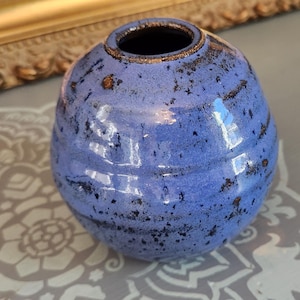 May include: A small, spherical ceramic vase with a vibrant periwinkle blue glaze. The surface features a textured, speckled finish with dark brown and black flecks throughout, and horizontal ridges circling the body of the vase.