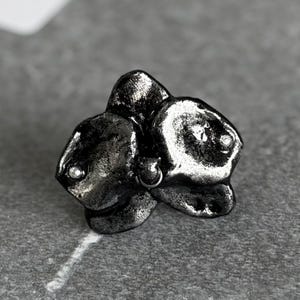 May include: A metallic silver-toned stud earring shaped like a three-petaled flower. The surface has a textured, uneven finish with a dark, oxidized appearance. Small, reflective circular accents are embedded within the petals of the flower.