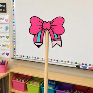 May include: A decorative pencil topper featuring a bright pink bow design with teal and black accents. The topper is attached to a plain, light-colored wooden stick, creating a handheld pointer or classroom accessory with a colorful graphic illustration.