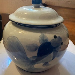 May include: A round ceramic jar with a lid featuring a blue and white hand-painted fish motif. The jar has a glossy white finish, a dark blue rim, and a small blue knob on the center of the lid. The fish design is rendered in shades of blue.