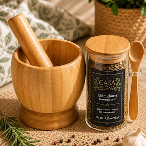 Peut inclure: Un mortier et un pilon en bois clair accompagnés d'un pot à épices en verre transparent avec un couvercle en bambou et une petite cuillère en bois. L'étiquette noire indique CASA ELENA Chimichurri Listo para usar Sabor auténtico casero Sin conservantes Net Wt. 3.25 oz (92 g).
