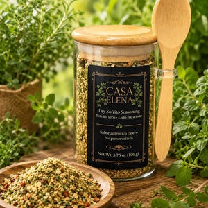 Może przedstawiać: Przezroczysty szklany słoik wypełniony suchą przyprawą Sofrito Casa Elena, z bambusową pokrywką i drewnianą łyżką przymocowaną z boku. Czarna etykieta zawiera tekst Casa Elena, Dry Sofrito Seasoning oraz Net Wt. 3.75 oz (106 g).