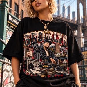 Puede incluir: Una camiseta negra de manga corta con un estampado gráfico que incluye la palabra BROOKLYN en letras grandes, un icono de corona dorada y un collage de una persona, un radiocasete, un disco de vinilo, una cinta de casete y una zapatilla.