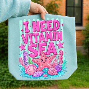 May include: A light blue pouch featuring a graphic print with the pink text I NEED VITAMIN SEA. The design includes a smiling pink starfish, two seahorses, various seashells, and coral elements set against a background of vertical light green stripes.