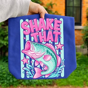 Pode incluir: Uma sacola azul com um gráfico de um peixe turquesa e rosa saltando da água. O texto SHAKE THAT está impresso em letras rosas grandes acima do peixe. O fundo inclui listras verticais azul-claras e pequenas estrelas cor-de-rosa.