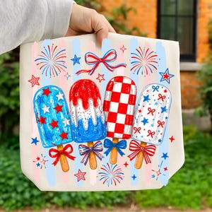 May include: A cream-colored tote bag featuring a graphic print of four popsicles in red, white, and blue. The design includes fireworks, stars, and bows, set against vertical pastel stripes. Each popsicle has a wooden stick and a decorative bow.