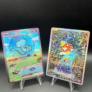 May include: Two holographic trading cards displayed on clear plastic stands. The left card features a blue Mew EX character with 180 HP, while the right card depicts a red Magikarp character with 30 HP set against a detailed, colorful forest waterfall background.
