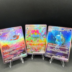 May include: Three holographic Pokemon trading cards displayed on clear plastic stands. The left card is Charizard ex with 330 HP, the center card is Venusaur ex with 340 HP, and the right card is Blastoise ex with 330 HP, each featuring colorful illustrations.