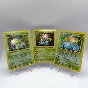 May include: Three holographic trading cards featuring Bulbasaur, Ivysaur, and Venusaur. Each card displays the character name, HP value, and specific attack text. The cards are arranged side-by-side, showcasing green borders and detailed illustrations of the creatures.