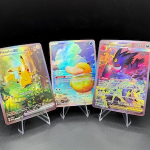 May include: Three holographic trading cards displayed on clear plastic stands. The left card features Pikachu with the text Thunderbolt, the center card shows Psyduck with the text Damp, and the right card depicts Mega Gengar EX with the text Shadowy Concealment.
