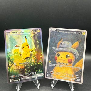 May include: Two Pokémon trading cards displayed on clear plastic stands. The left card is Pikachu ex with 200 HP and the Thunderbolt attack. The right card is Pikachu with Grey Felt Hat, 60 HP, and the Pika-Portrait attack, featuring an artistic style.