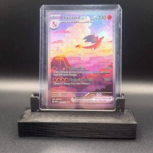 May include: A Charizard ex trading card in a clear protective sleeve, displayed on a black 3D-printed stand. The card features a vibrant illustration of a dragon flying over a volcanic landscape, with text including Brave Wing and Explosive Vortex.