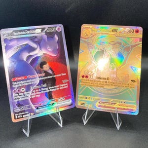 May include: Two holographic trading cards displayed on clear plastic stands. The left card features a purple Mewtwo character with 280 HP, while the right card shows a gold-toned Mega Charizard X character with 360 HP and fire-type symbols.