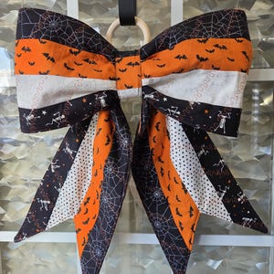 May include: A large fabric bow featuring a patchwork of Halloween-themed patterns including black spiderwebs on a dark background, orange fabric with black bats, white fabric with black dots, and black fabric with white skeletons and stars.