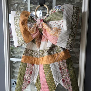 May include: A large, quilted fabric bow in shades of sage green, soft pink, and cream, featuring various floral and text-printed patterns. The bow is constructed from multiple stitched fabric strips and is suspended from a circular wooden ring.