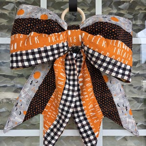 May include: A large decorative fabric bow featuring a mix of Halloween-themed patterns including orange pumpkins, black spiderwebs, and white skeletons on gray, black, and orange backgrounds with text reading EEEK and SCARY TREATS in white.