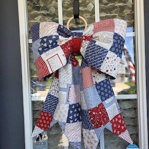 May include: A large decorative bow made of fabric with a patchwork design in red, white, and blue. The fabric features various patterns including text listing US states, small stars, and geometric shapes. The bow is attached to a light wooden ring.
