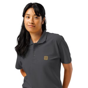 Könnte beinhalten: Ein dunkel anthrazitfarbenes Kurzarm-Poloshirt mit klassischem Kragen, einer Knopfleiste mit drei Knöpfen und einem kleinen quadratischen beigen Aufnäher auf der linken Brust mit einem dunkelbraunen, kreisförmigen Logo.