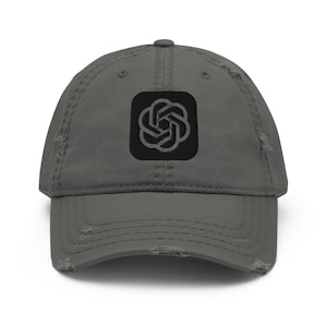 Puede incluir: Una gorra de béisbol gris carbón con aspecto desgastado y un parche cuadrado negro en el panel frontal. El parche muestra un logotipo geométrico circular de color gris oscuro formado por seis bucles entrelazados en forma de flor.