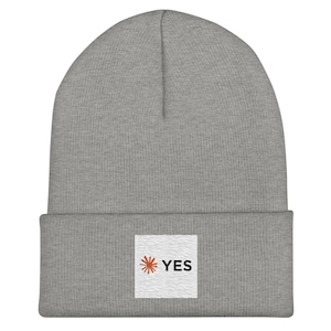 May include: A heather grey knit beanie with a cuffed brim. Centered on the cuff is a square white patch featuring a pattern of thin wavy lines, a small orange sunburst icon, and the word YES printed in bold black capital letters.