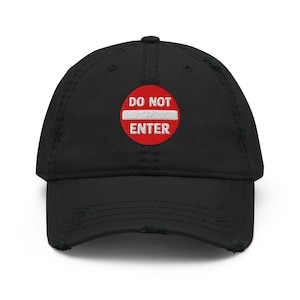 Könnte beinhalten: Eine schwarze, abgenutzte Baseballkappe mit einem gestickten, kreisförmigen roten Aufnäher auf der Vorderseite. Der Aufnäher zeigt einen weißen horizontalen Balken und den weißen Text DO NOT ENTER, ähnlich einem Verkehrsschild.