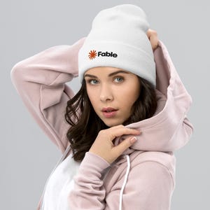 Op de afbeelding: Een witte muts met omslag en een geborduurd logo op de voorzijde. Het logo bestaat uit een oranje zon-icoon links van de zwarte tekst "Fable". De muts is gemaakt van een geribbelde gebreide stof en heeft een omgeslagen boord.