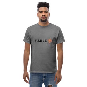 May include: A charcoal gray short-sleeve t-shirt featuring the word FABLE printed in black capital letters on the chest, followed by a small, eight-pointed star-like graphic in a muted orange color. The shirt has a crew neckline and a standard fit.