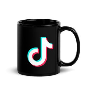 Könnte beinhalten: Eine schwarze Keramiktasse mit dem TikTok-Logo auf der Vorderseite. Das Logo ist ein weißes Notensymbol mit cyanfarbenen und magentafarbenen Farbverschiebungen an den Rändern, die einen Schichteffekt auf dem schwarzen Hintergrund der Tasse erzeugen.