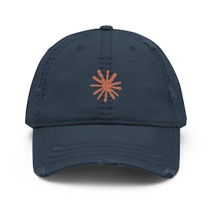 Könnte beinhalten: Eine marineblaue Baseballkappe mit abgenutztem, distressed Finish. Auf der Vorderseite befindet sich eine pfirsichfarbene Stickerei in Form eines Sonnen- oder Sternenstrahls mit mehreren strahlenförmigen Linien in der Mitte.