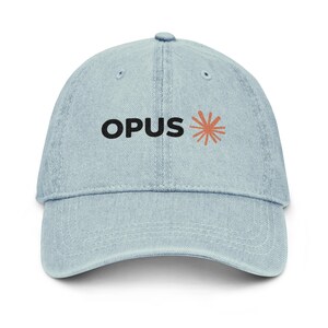 Pode incluir: Um boné de beisebol de ganga azul claro com a palavra OPUS bordada em letras maiúsculas pretas, seguida de um gráfico estilizado de explosão solar laranja no painel frontal. O boné tem uma aba curva e um design estruturado de seis painéis.