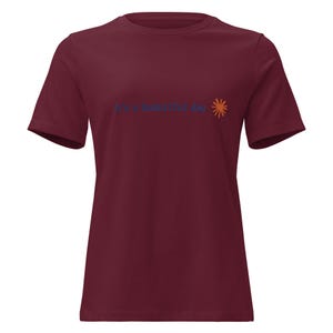 May include: A maroon short-sleeved t-shirt featuring the text "it's a beautiful day" printed in dark blue letters on the chest, accompanied by a small, stylised orange sun icon positioned to the right of the text.