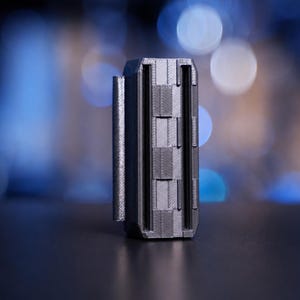May include: A dark gray, rectangular, 3D-printed object featuring a vertical hinge mechanism on one side and a series of stacked, textured segments along the front face. The item has a matte finish with visible layer lines from the printing process.