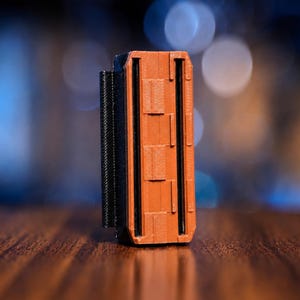 May include: A rectangular 3D printed object featuring a textured orange front panel with a vertical brick-like pattern and a black side frame. The structure includes two parallel vertical slots running along the length of the orange face.