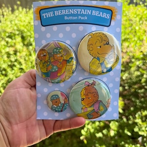 Può includere: Un set di quattro spille rotonde con i personaggi di The Berenstain Bears, montate su un cartoncino azzurro a pois bianchi. Il cartoncino riporta in alto la scritta THE BERENSTAIN BEARS Button Pack all'interno di un banner blu.