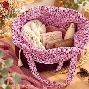 Peut inclure: Un sac fourre-tout en velours côtelé violet foncé avec une doublure intérieure à motif d'éventails magenta et blanc. Le sac possède une fermeture à cordon avec des liens blancs et des perles en bois, contenant une écharpe en tricot crème.