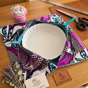 Peut inclure: Un porte-plat en tissu arborant un motif floral vibrant violet, blanc et bleu sarcelle avec une doublure intérieure unie bleu sarcelle. Il contient un plat de cuisson rond en céramique blanche à bord cannelé. Une étiquette en cuir indique RoseKiwi Créations.