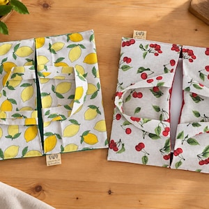 Peut inclure: Deux sacs en tissu avec poignées, l'un présentant un motif de citrons jaunes sur fond blanc et l'autre un motif de cerises rouges sur fond gris clair. Chaque sac possède une petite étiquette rectangulaire fixée sur le bord inférieur.