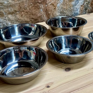 May include: A set of five polished stainless steel mixing bowls of varying sizes. Each bowl features a smooth, reflective surface, a rounded base, and a wide, slightly flared rim. The bowls are arranged in a cluster, showcasing their metallic finish.