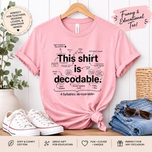 Op de afbeelding: Een lichtroze T-shirt met korte mouwen en zwarte tekst die This shirt is decodable leest. Het ontwerp bevat diverse taalkundige annotaties zoals High-Frequency Word, Digraph, Prefix, Schwa Sound en Suffix, met een uitsplitsing van de lettergrepen.