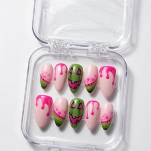 May include: A set of ten almond-shaped press-on nails in a clear plastic case. The designs include pink drip patterns, watermelon-inspired segments with green rinds and black seeds, and green jack-o'-lantern faces with pink carved features.