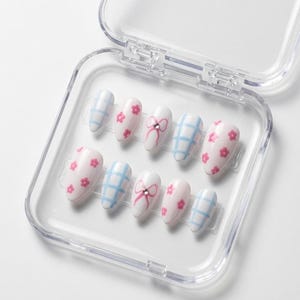 Op de afbeelding: Een set van tien kunstnagels in een doorzichtig plastic doosje. De nagels hebben een witte basis met diverse ontwerpen, waaronder lichtblauwe ruitpatronen, kleine roze bloemetjes en een roze strikmotief met een klein doorzichtig steentje.