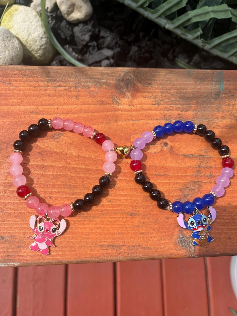 May include: Two beaded bracelets connected by a gold heart-shaped magnetic clasp. The left bracelet features pink, red, and black beads with a pink character charm. The right bracelet features blue, red, black, and light purple beads with a blue character charm.