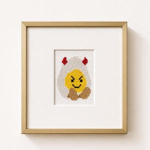 May include: A cross-stitch embroidery in a gold frame featuring a yellow circular face with a black smile and eyes, wearing a white hood with two small red horns and brown accents at the bottom, set against a white background within the frame.