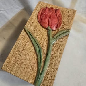 May include: A rectangular wooden plaque featuring a hand-carved and painted relief of a single tulip. The flower head is painted in shades of red, while the stem and leaves are green. The background wood has a textured, natural grain finish.