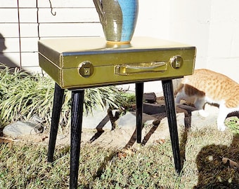 May include: A gold-coloured rectangular suitcase repurposed as a small table with four tapered black wooden legs. A ceramic pitcher with a blue and brown glazed finish sits on top of the suitcase. The object is positioned outdoors on grass.