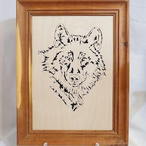 Könnte beinhalten: Ein rechteckiger Holzrahmen mit einer hellen Holzplatte, die eine schwarze Brandmalerei eines Wolfskopfes zeigt. Der Wolf ist von vorne dargestellt, mit detaillierter Fellstruktur und spitzen Ohren, zentriert im Rahmen.