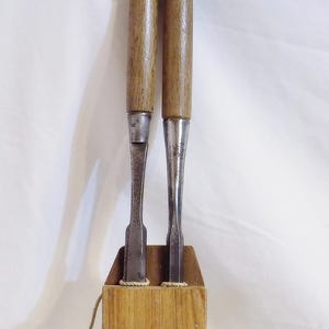 May include: Two metal woodworking chisels with light brown wooden handles and silver-colored metal ferrules, standing upright in a small, rectangular wooden block holder with a thin cord attached to the side and a copper penny nearby.