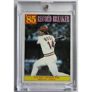 Pode incluir: Um cartão de beisebol de Pete Rose de 1985 em um estojo protetor de plástico transparente. O cartão mostra um jogador com uniforme branco rebatendo, com o texto '85 RECORD BREAKER' no topo e 'PETE ROSE, CINCINNATI REDS Most Hits, Lifetime' na parte inferior.